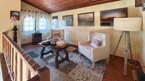 Un salon chaleureux avec un plafond en bois, un canapé, un fauteuil, une table basse et une lampe. Il y a des fenêtres avec des rideaux et des tableaux encadrés accrochés au mur.
