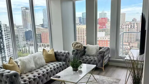 Ein modernes Wohnzimmer mit einem grauen, gesteppten Sofa, einem weißen Couchtisch und Blick auf die Stadt durch große Fenster.
