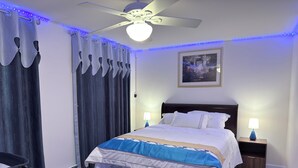 Ein ordentlich eingerichtetes Schlafzimmer mit Bett, Deckenventilator, Nachttisch mit Lampe und einem Wandbild.