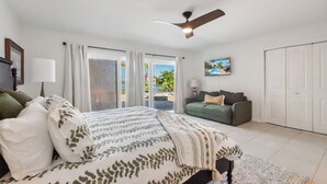 Una camera da letto con un letto, un divano verde, una ventola a soffitto e vista su una piscina e palme attraverso la finestra.