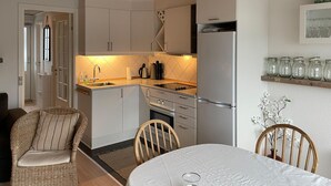 Cocina compacta con armarios blancos, un lavamanos de acero inoxidable y un refrigerador. Hay una mesa de comedor con sillas y una pequeña planta en maceta.