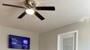 Un salon avec un canapé gris, un ventilateur de plafond, un téléviseur fixé au mur et une porte.