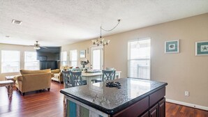A spacious living room with a large wooden floor, a beige sofa, and a dining area with a kitchen island. - Easy Gulf-View Retreat w/ Big Deck, Outdoor Shower & Pet Friendly (Bolivar Peninsula)