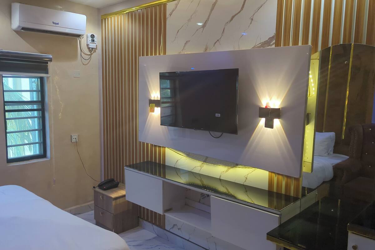 Una habitación de hotel moderna con un televisor de pantalla plana grande montado en una pared de mármol, una mesita de noche con lámpara, una pequeña mesita de noche con teléfono y una ventana con persianas.