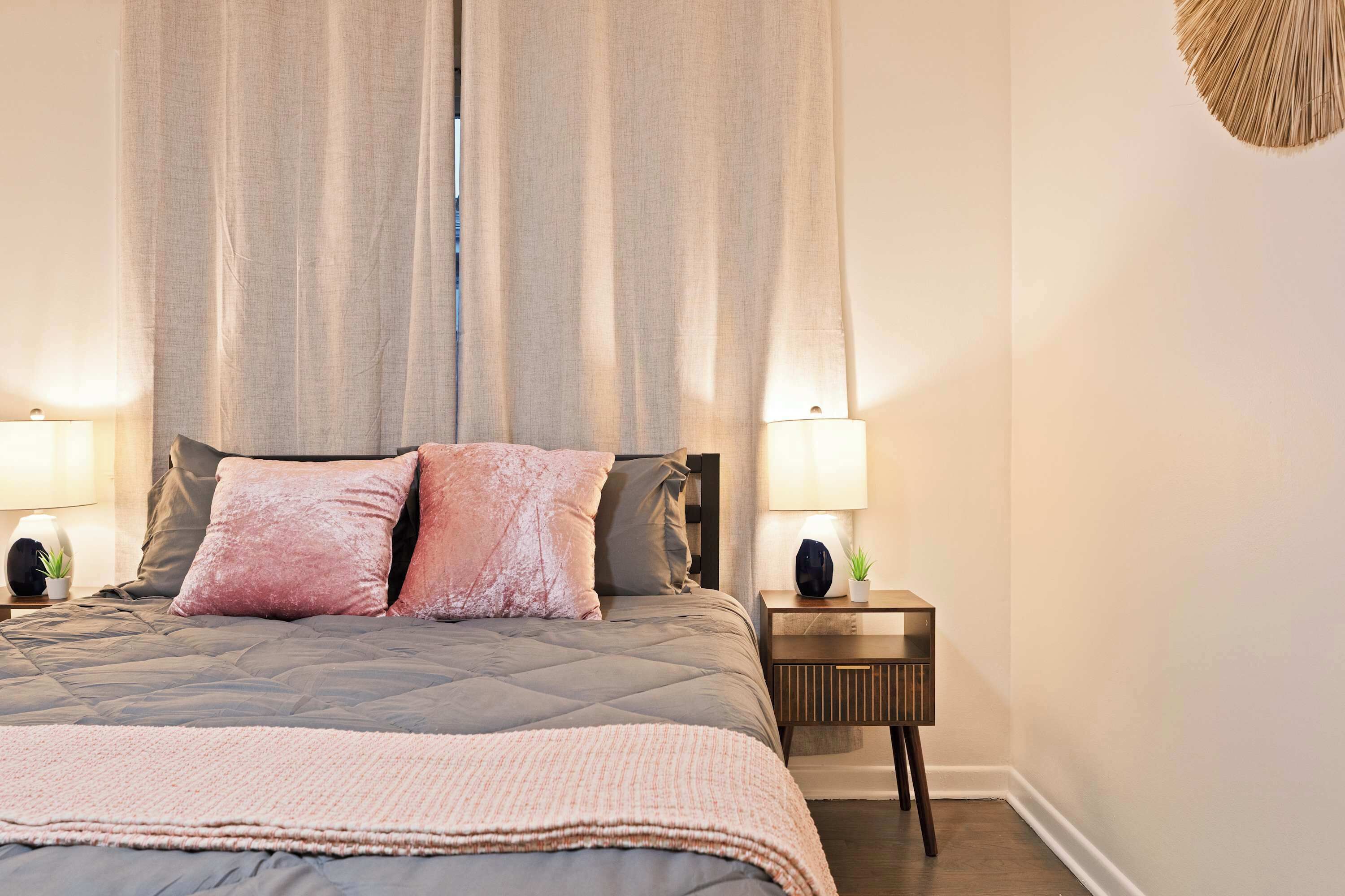 A bedroom with a large bed, two bedside lamps, a nightstand with a plant, and a window with curtains.