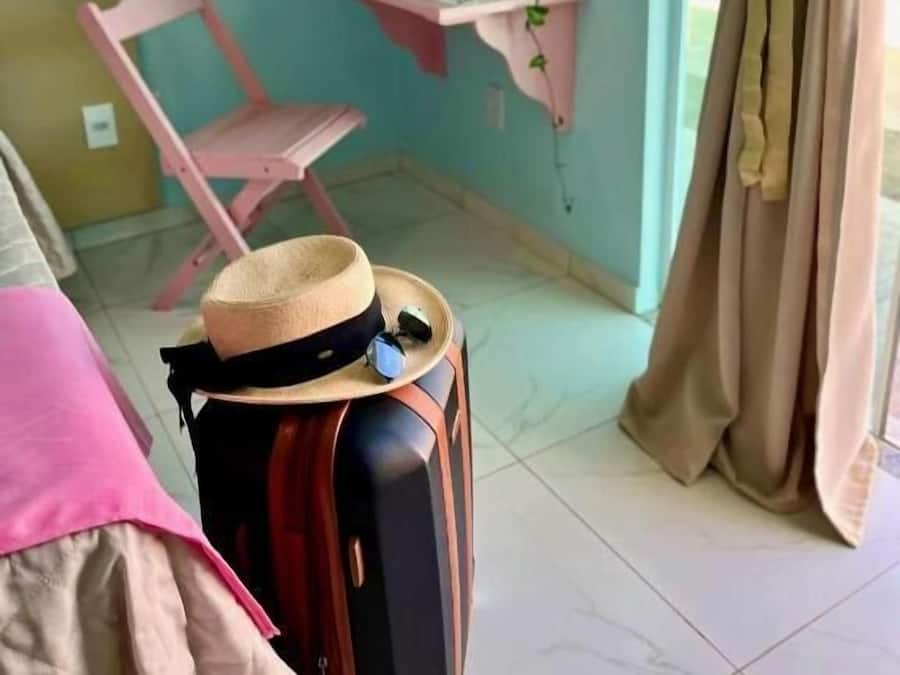 Una habitación con una silla rosa, una pequeña mesa con una planta y una maleta. La pared es color verde azulado y tiene los textos 'villa monale' y 'desayuno'.