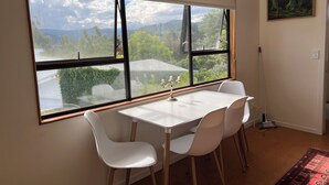 Een eethoek met een tafel en stoelen, een raam met uitzicht op groen en bergen, en een schilderij aan de muur.