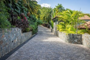 Un sentier pavé de pierres, bordé de murs en pierre et d’une végétation tropicale luxuriante, mène à un bâtiment surmonté d’un toit en tuiles.