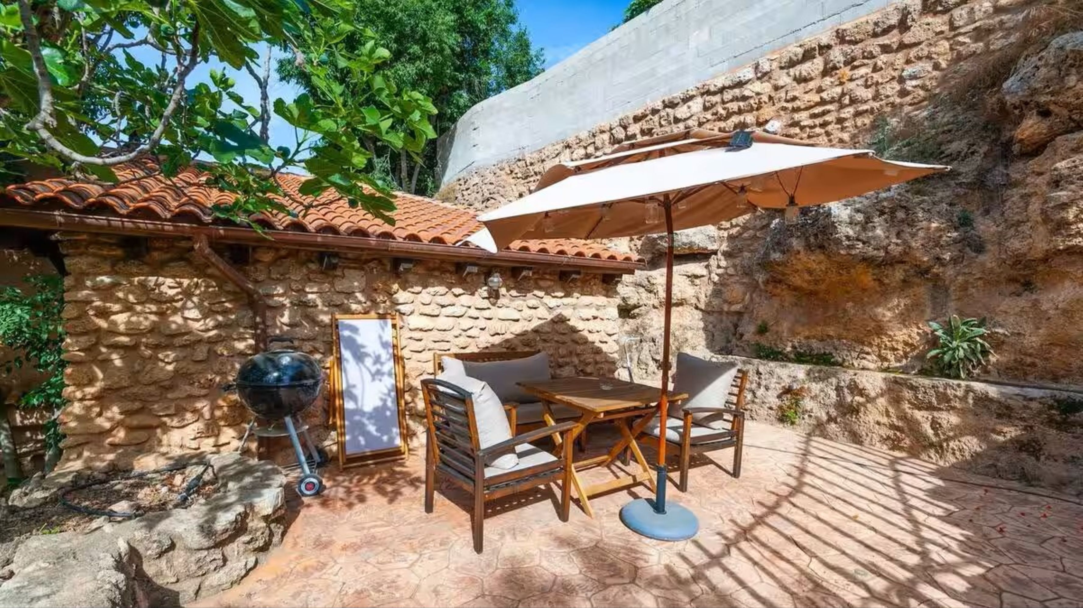 An outdoor patio with stone walls, a tiled roof, and a table with chairs and an umbrella.