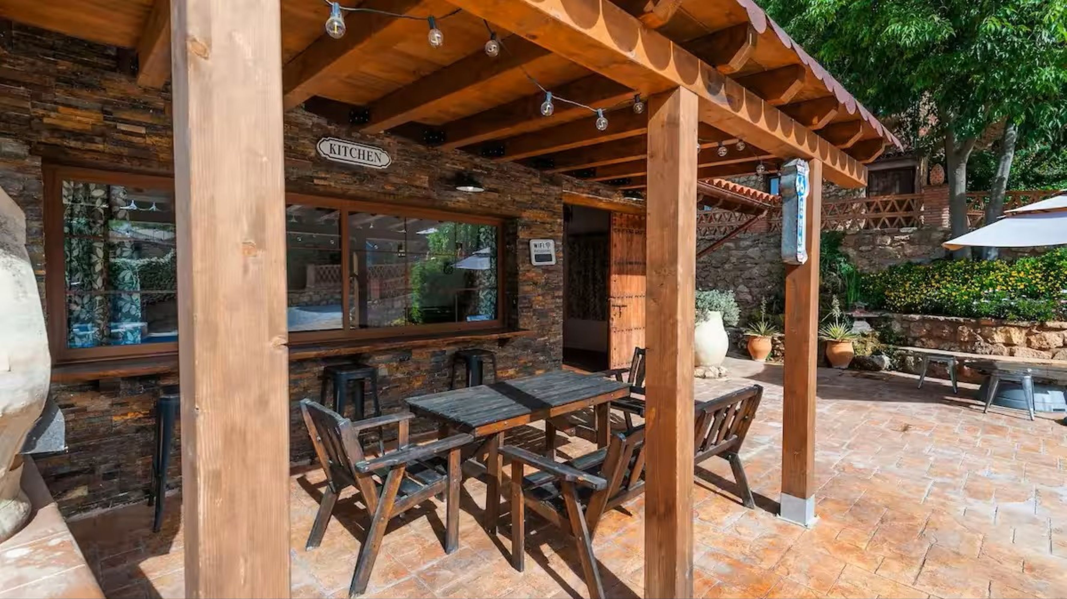 An outdoor patio with wooden tables and chairs, a stone wall, and a sign indicating a kitchen area.