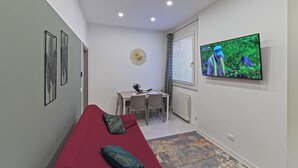 A compact living space with a red sofa, a dining table with chairs, a TV mounted on the wall, and a ceiling fan. - Paint Venice House (Venezia)