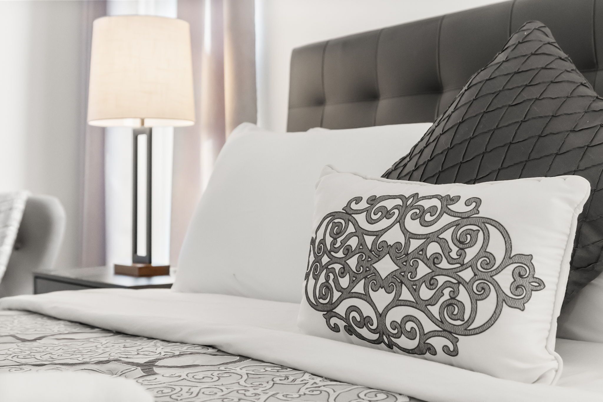 Een net opgemaakt bed met een bedspreide met een patroon, een decoratief kussen met een ingewikkeld ontwerp en een moderne lamp ernaast.