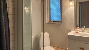 Un baño con cabina de ducha de vidrio, inodoro blanco, pequeña ventana con un diseño azul, lavamanos con espejo y una botella de jabón para manos.