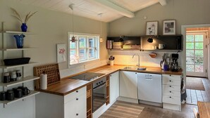 Una cocina moderna con armarios blancos, encimera de madera y horno empotrado. Hay una ventana que proporciona luz natural, y la cocina está equipada con un lavamanos y varios utensilios.