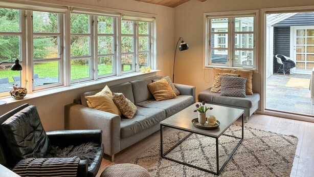 Una sala de estar acogedora con un sofá gris, un sillón de cuero negro y una mesa de centro. Se observan amplias ventanas con marcos blancos, una lámpara de pie negra y vistas a un jardín exterior.