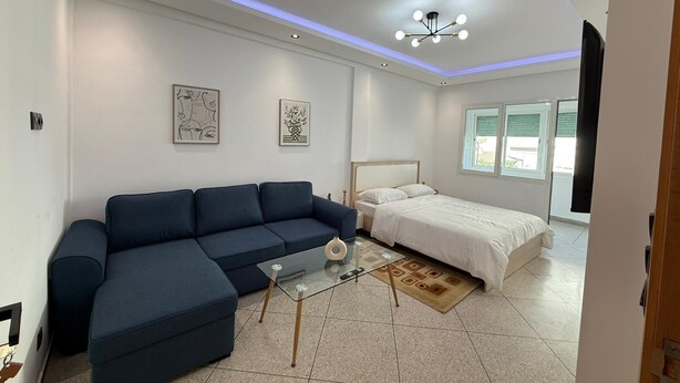 Une chambre moderne avec un canapé bleu, une table basse en verre, un lit recouvert d’une couette blanche et deux œuvres d’art encadrées accrochées au mur.