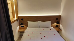 Ein ordentlich bezogenes Bett mit weißen Leinentüchern und verstreuten Rosenblättern. Zwei Nachttische, jeder mit einer schwarzen Lampe. Das Kopfteil ist aus Holz, und der Raum hat ein schlichtes, aufgeräumtes Design.