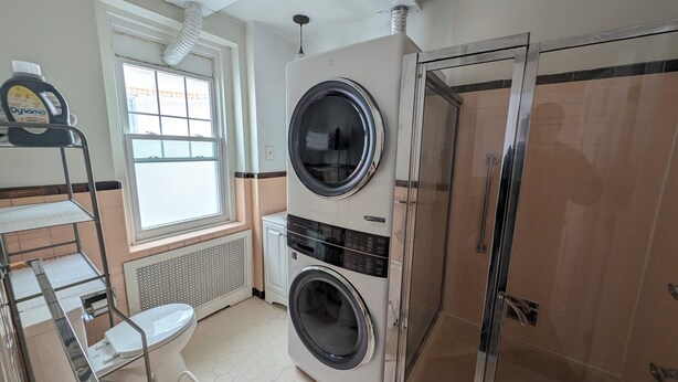 A laundry room with two stacked front-loading washing machines, a window, a shelf with cleaning supplies, and a white toilet.