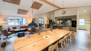 Een ruime woonkamer met een lange houten tafel, comfortabele zittingen en een keuken op de achtergrond.