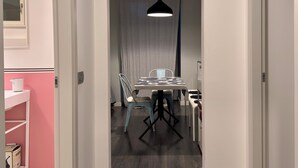Un interior modern amb una zona de menjador que inclou taula i cadires, una làmpada penjant, i una vista cap a una altra habitació amb finestra i cortines.