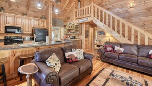 A spacious wooden cabin interior with a kitchen, dining area, and living room furnished with leather sofas and a staircase. - Black Bear Ridge (Blairsville)