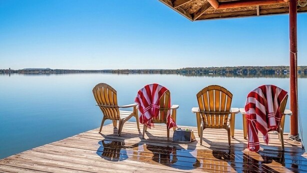 A wooden deck with chairs and a table, overlooking a calm lake and distant hills. - Stunning Views, Open Water, Hot Tub, 5 BR, 4 BA (Granbury)