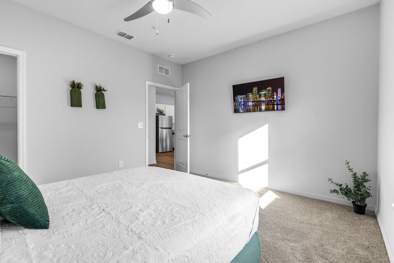 A bedroom with a bed, a ceiling fan, a cityscape painting, and a view of the kitchen.
