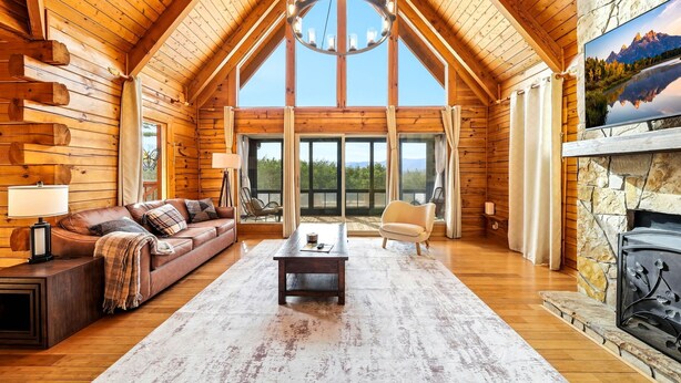 Ein Interieur einer Blockhütte mit Holzbalkendecke, Kamin, einer Couch, einem Couchtisch und einem großen Fenster mit Blick ins Freie.