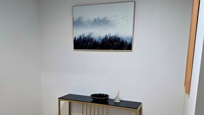 A room with a wooden floor, a black console table with a gold frame, a black stool, and a wall-mounted painting of a forest scene.