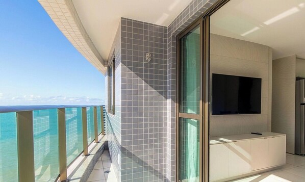 A balcony with a glass railing, a flat-screen TV mounted on the wall, and a tiled wall. - Apto w/ Balcony Beachfront in Boa Viagem Cpf0902 (Recife)