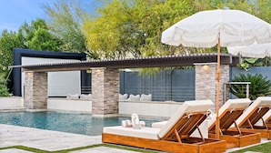 A pool area with lounge chairs and umbrellas, surrounded by a stone wall and greenery. - Luxury Resort Oasis : Pool, Pickleball, Sauna, Spa,     Sleeps 14 Comfortably  (Phoenix)
