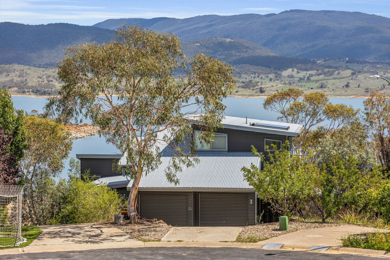 A modern house with a garage, surrounded by trees and a lake, with mountains in the background.