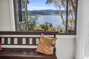 A wooden bench with a cushion, a window with a view of a lake and mountains, and a white wall. - Lakeview Lodge | East Jindabyne | Lakefront | 3BR (East Jindabyne)