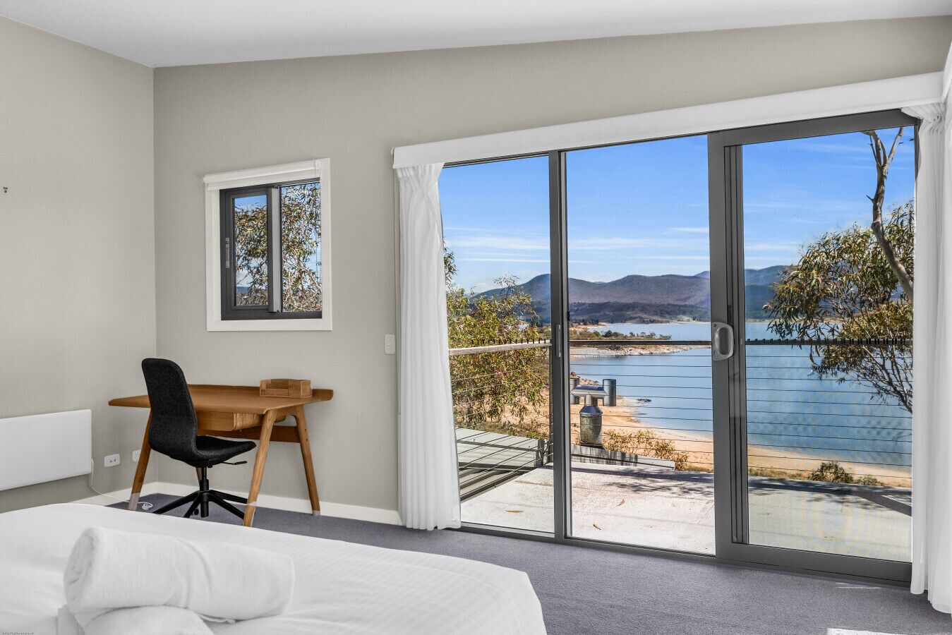 Une chambre avec un lit, un bureau et une chaise, ainsi qu’une grande porte-fenêtre coulissante donnant sur un balcon avec vue sur un lac et des montagnes.