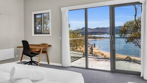 Un dormitorio con cama, escritorio y silla, y un amplio ventanal corredero a un balcón con vistas a un lago y montañas.