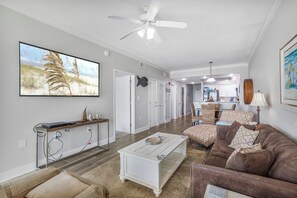 A spacious living room with a large flat-screen TV, a ceiling fan, a brown leather sofa, and a coffee table. - Gulf Front 2 Bedroom Condo in Gulf Shores - Crystal Shores Unit 404 (Gulf Shores)