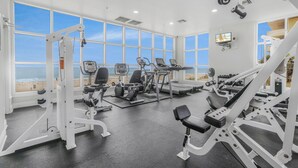 Une salle de sport équipée de divers appareils d’exercice, dotée de grandes fenêtres et offrant une vue sur la plage.
