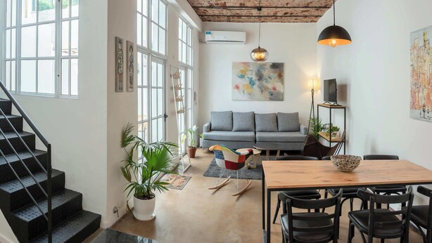 Een ruime woonkamer met een grijze bank, een houten eettafel en zwarte stoelen. Links bevindt zich een trap, een groot raam en een airconditioning op de muur.