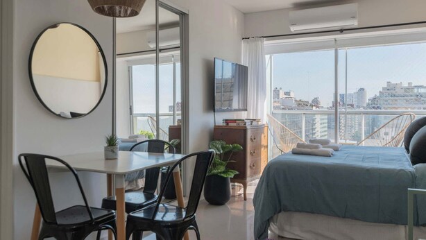 Une chambre moderne avec un grand lit, une table à manger avec des chaises, un téléviseur et un balcon offrant une vue sur la ville.