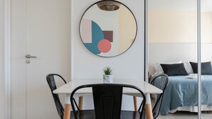 Une chambre d’hôtel moderne avec un coin repas comprenant une table blanche et des chaises noires, une œuvre d’art murale ronde, une petite plante en pot et un lit avec une couvre-lit bleue et des oreillers noirs.