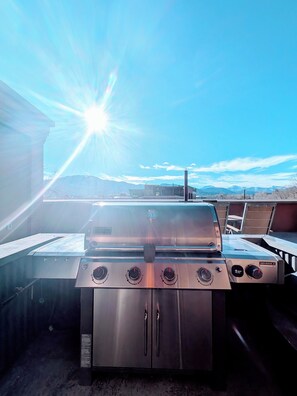 Ein Edelstahl-Grill für den Außenbereich mit vier Brennern und einem Schornstein, vor einem strahlend blauen Himmel mit Bergen im Hintergrund.