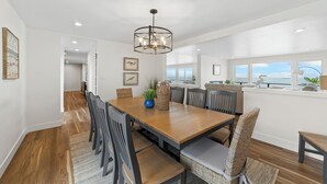 A spacious dining area with a wooden table and chairs, a chandelier, and a view of the ocean through large windows. - Hot tub on the Beach with Super-Fast Wi-Fi (Oak Harbor)