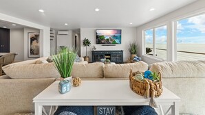 A modern living room with a beige sofa, a white coffee table with decorative items, a flat-screen TV on the wall, and a view of the ocean through large windows. - Hot tub on the Beach with Super-Fast Wi-Fi (Oak Harbor)