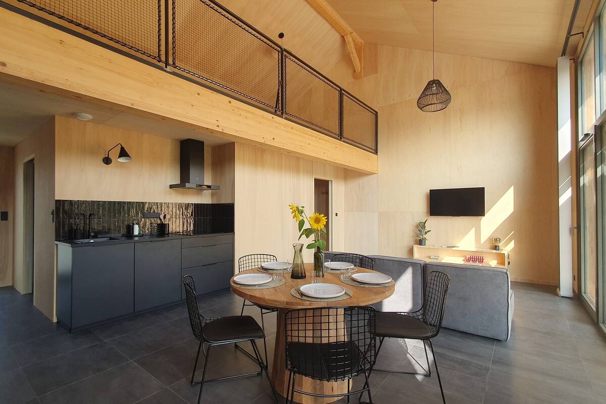 A modern kitchen with a dining area, featuring a wooden ceiling, large windows, and a central island with chairs.