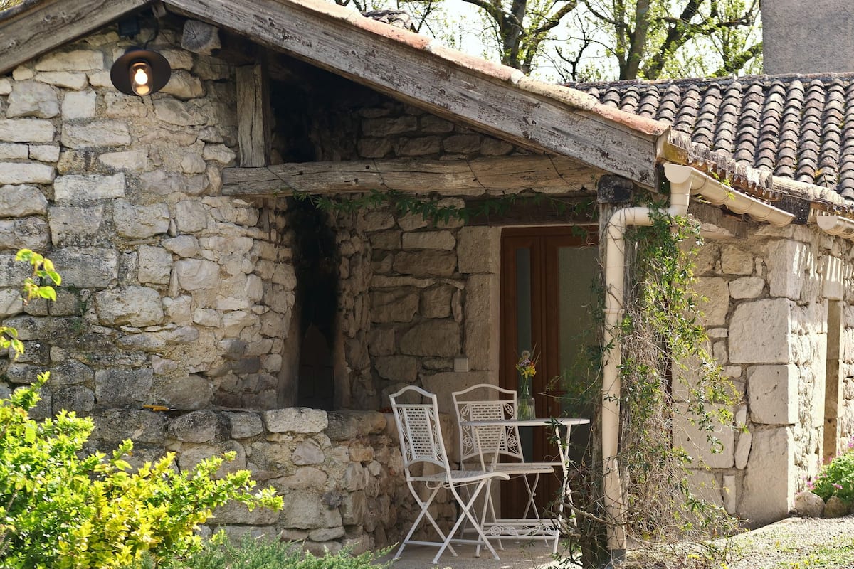 A stone building with a sloped roof, a wooden door, and a small outdoor seating area with two chairs and a table.
