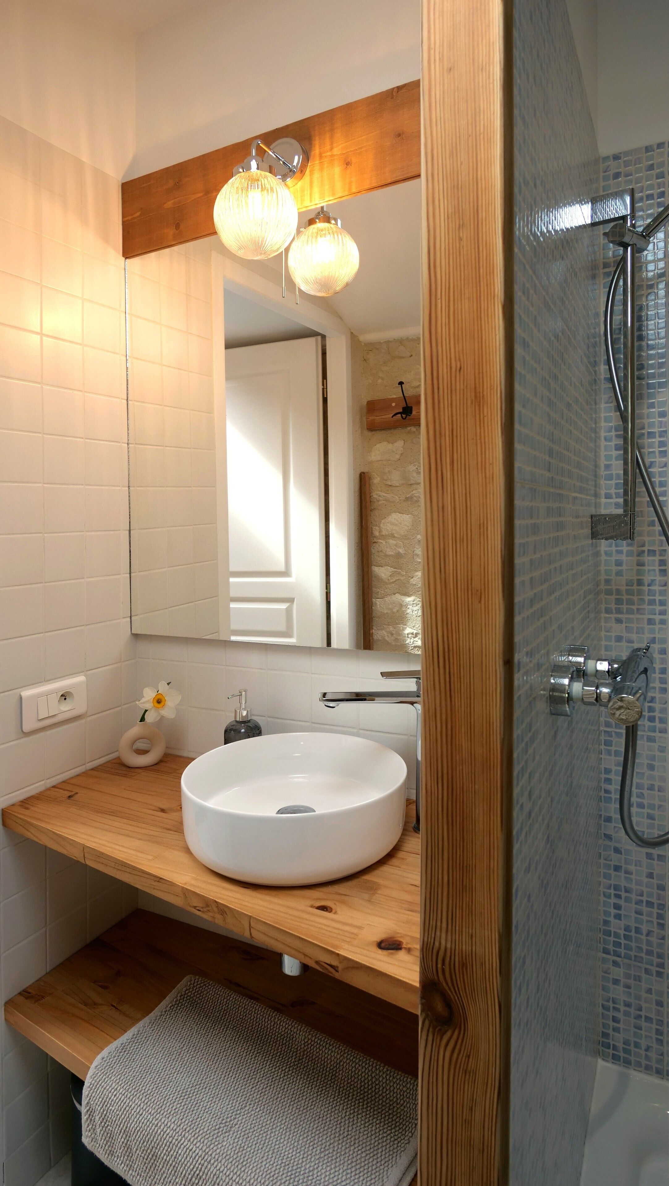 Un baño con tocador de madera, un lavamanos blanco, un espejo con marco de madera y una zona de ducha con mosaicos azules.