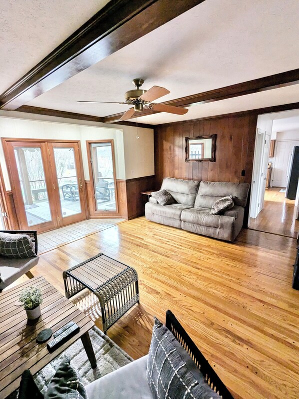 A spacious living area with a wooden floor, a grey sofa, a ceiling fan, and a view through glass doors to an outdoor patio.