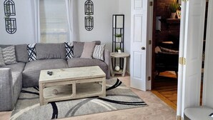 Ein Wohnzimmer mit grauem Sofa, einem Holztisch und einem Teppich mit Muster. Eine Tür führt zu einem Bereich mit Kamin aus Ziegelsteinen.