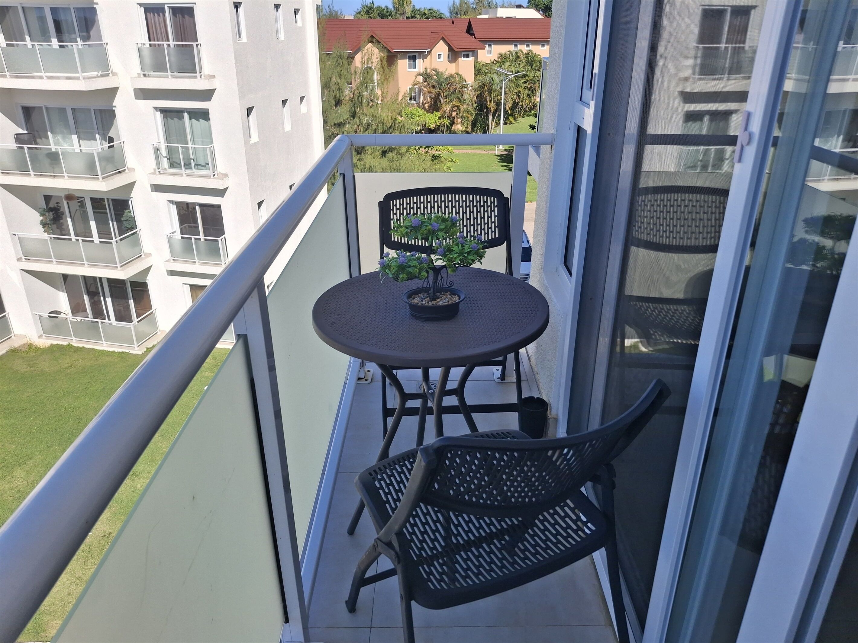 Un balcon avec une table ronde et deux chaises, un pot de fleurs et une vue sur les bâtiments voisins et la verdure.