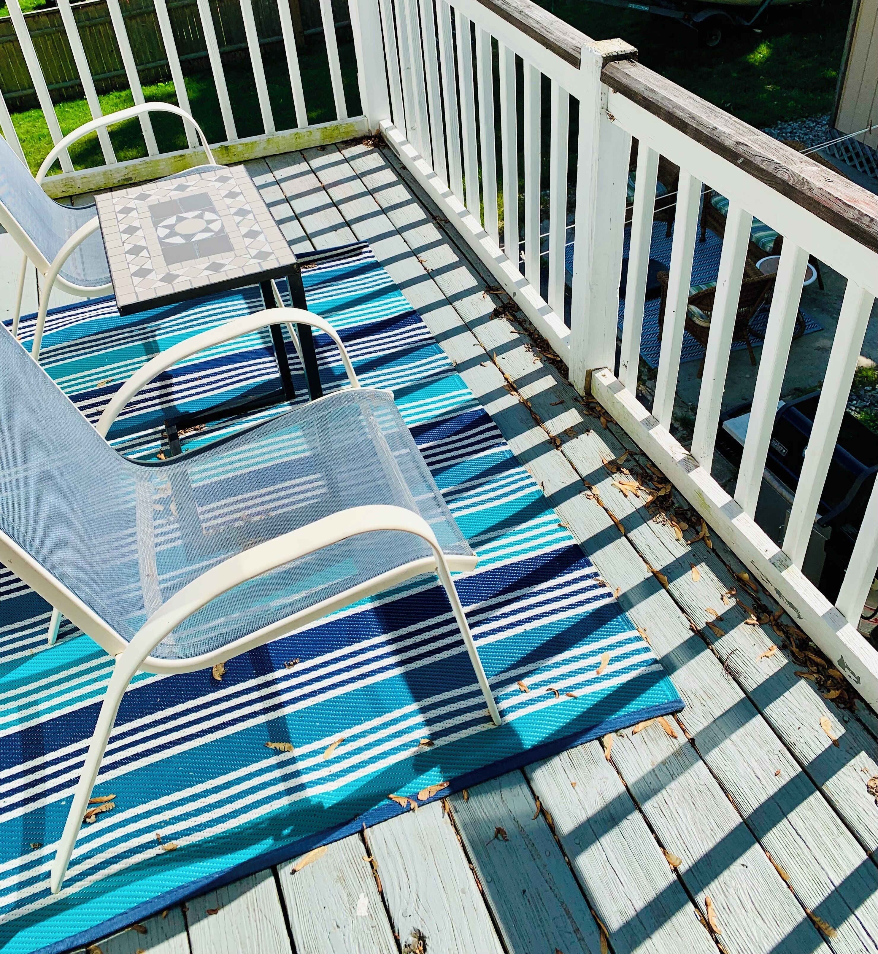 Una terraza de madera con un tapete azul y blanco a rayas, dos sillas metálicas y una mesita.